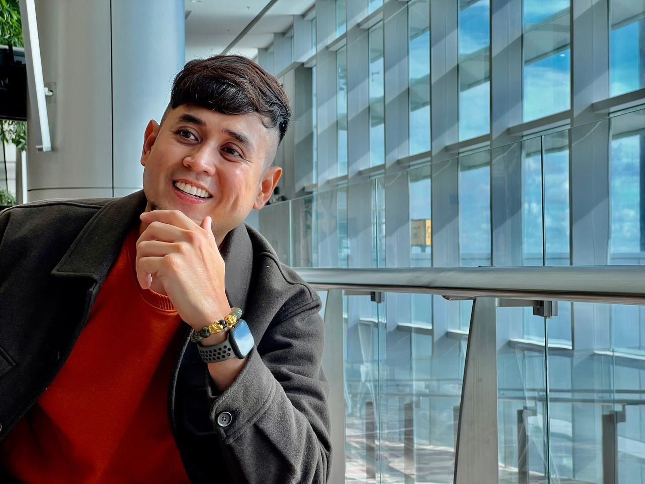 A man with short black hair smiles and poses indoors, wearing a coat.